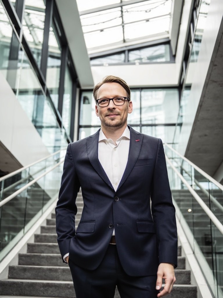 René Schröder auf der Treppe im gläsernen Atrium eines Bürogebäudes, dunkelblauer Anzug — Porträt zum Beitrag über Projektverzögerungen